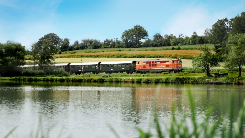 Reblaus Express, © NÖVOG-weinfranz Ein Zug fährt entlang eines Flusses durch eine grüne Landschaft unter blauem Himmel.