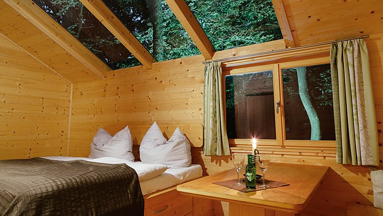 Innenansicht einer Holzhütte mit Bett, Tisch und Kerze, durch Fenster und Dachfenster sind Bäume sichtbar.