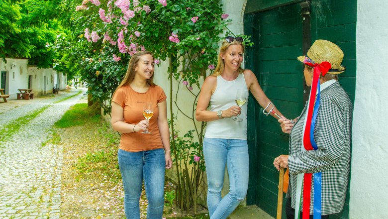 Drei Personen stehen vor einem grünen Tor, zwei halten Weingläser, eine trägt einen Strohhut mit bunten Bändern.