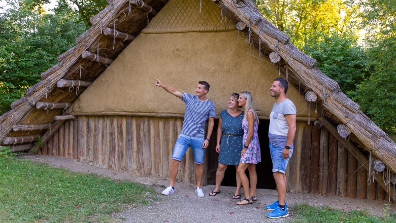 Vier Personen stehen vor einem rekonstruierten prähistorischen Haus im Freien.